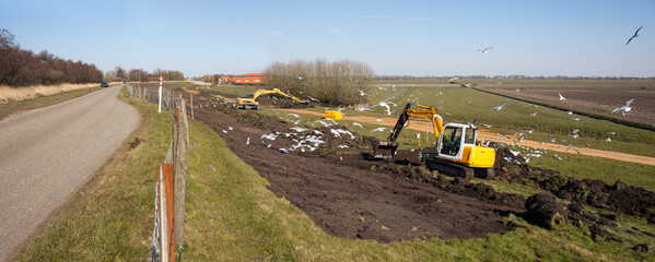 A crane with backhoe is removing grass and topsoil from a dike. Gulls are having a feast and surround the machine. 