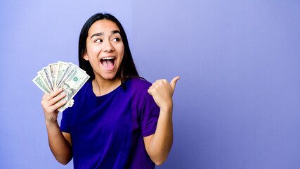 Young asian woman holding money isolated on purple background points with thumb finger away, laughing and carefree.