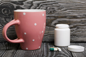 Container with pills and a mug of water. The lid of the container and several tablets are next to each other. On pine planks in black.