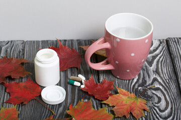 A container with pills, a mug of water and autumn maple leaves. The lid of the container and several tablets are next to each other. On pine planks in black.