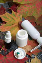 Pill container, inhaler and nasal drops. The lid of the container and several tablets are next to each other. Among the autumn maple leaves on the black pine boards.