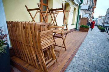 Stack of folded wooden chairs and tables on summer terrace of street cafe. Restaurants closing the summer season. Street cafe before opening. Folded chairs in closed restaurant.
