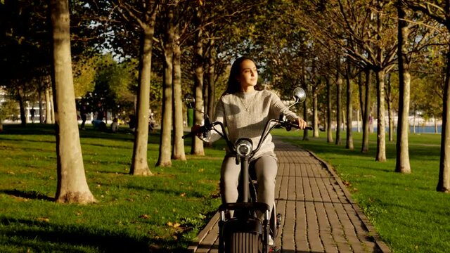 Young girl on electric scooter is riding through publick park at sunny morning, front view steadicam shot. Ecological electro transport concept.