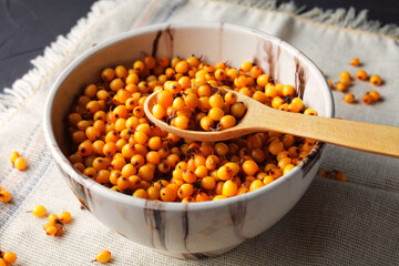 Fresh sea buckthorn in a clay bowl with a wooden spoon on a dark table, close-up.