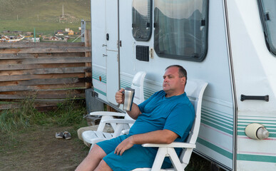 man is relaxing with a travel trailer RV. person on vacation with a mug in his hands. the concept of outdoor recreation. recreation water, long-distance travel and ecotourism. 