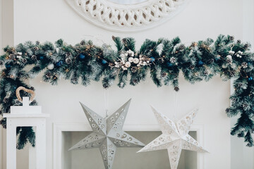 View over decorated artificial fireplace with decorations inside. Handmade paper stars, candles inside. Beautiful coniferous wreath on the top of fireplace. Decorative round mirror over the fireplace.