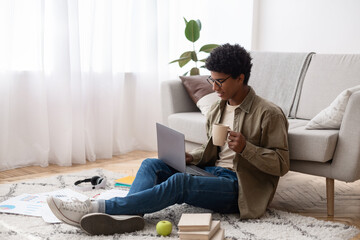 Attractive black teenager using laptop and drinking coffee during his online studies at home, copy space