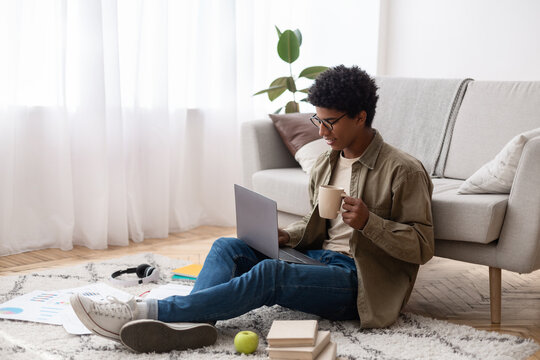 Attractive black teenager using laptop and drinking coffee during his online studies at home, copy space