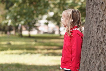 Fototapeta premium Little girl in a red jacket stands near a large tree in the park