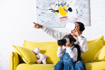 Mother pointing with finger and daughter using virtual reality headsets at home