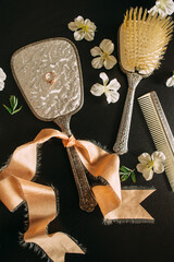 Vintage toiletries. Silver colors. Comb, mirror, golden ring, peach ribbon, white flowers, brush on black background