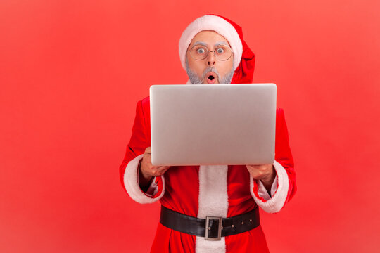 Shocked surprised santa claus peeping behind laptop, looking at camera with big wondered eyes. Indoor studio shot isolated on red background