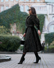 Fototapeta premium Stylish woman in black long coat with belt, skirt, tights, sunglasses and high heels walking at city street. Brunette caucasian girl with long wavy hair outdoor portrait