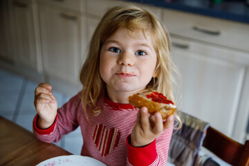 Cute funny toddler girl eats sweet bun for breakfast. Happy child eating bread roll with strawberry...