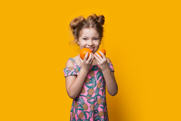 Charming blonde girl tasting sliced oranges posing in a dress on a yellow studio wall