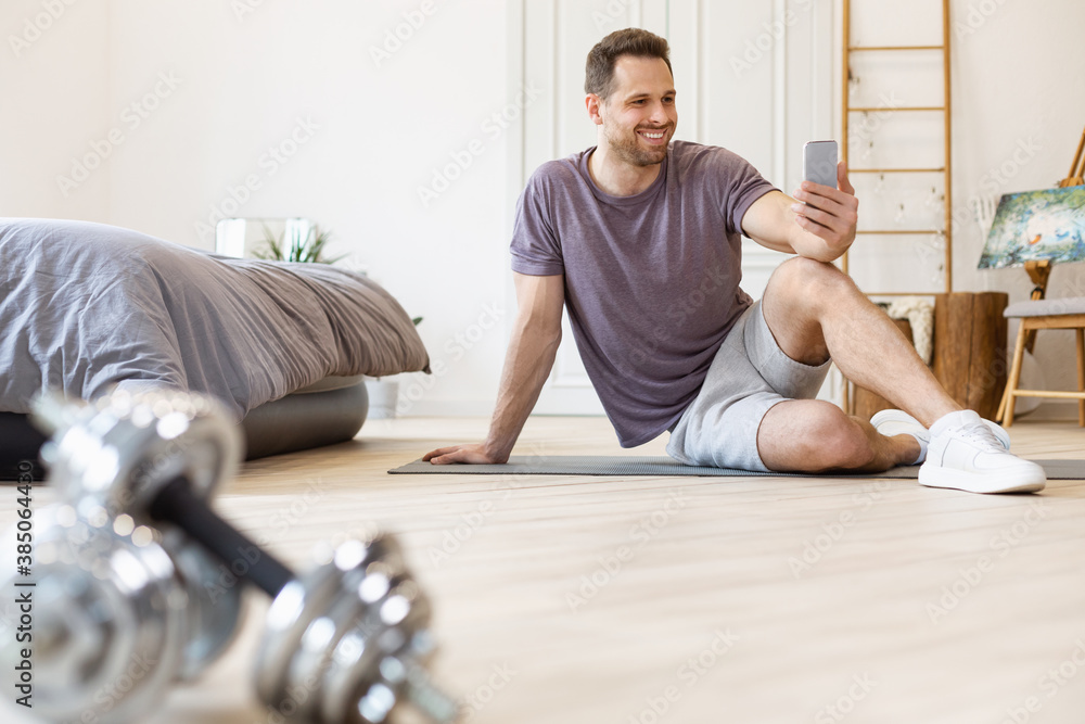 Poster man training using smartphone exercising sitting on mat at home