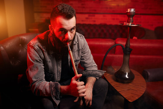 cheerful bearded successful guy in a dark room. close up side view photo. young man smokes a hookah while sitting. red smoke