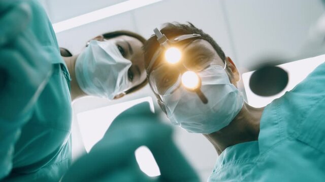 Surgeon with nurse are looking down at camera. Handsome young doctor in surgical mask is holding tools. Surgeons are standing above of the patient before surgery