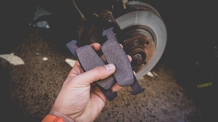 A man's hand with new brake pads on the background of a car without a wheel. Service work on replacing the front brake pads. - obrazy, fototapety, plakaty