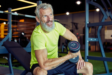 Portrait of a strong mature man in sportswear lifting heavy dumbbells and pumping his biceps while...