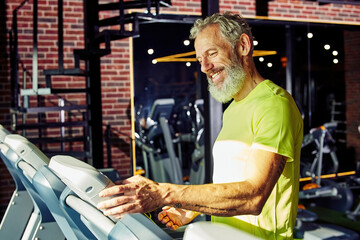 Obraz na płótnie Canvas Side view of a happy middle aged athletic man in sportswear adjusting speed on a treadmill while having cardio workout in a gym or sport club