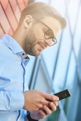 Side view of a young handsome businessman wearing wireless earphones using his smartphone while leaning against brick wall outdoors