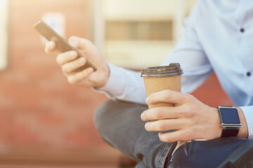 Cropped shot of a businessman in blue shirt using smartphone and drinking coffee while relaxing on the bench outdoors