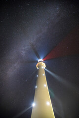 Night scene with the historic lighthouse in the Milky Way background with colorful navigation lights and white interior lights emitting mystic light beams into foggy autumn air