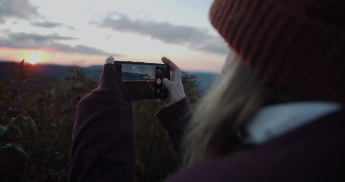 Young woman taking a picture on a phone of a sunset in the blue ridge mountains