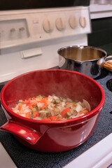 Steam rises from gumbo, a southern specialty dish with  vegetables, okra, Abelmoschus esculentus, bell pepper, Capsicum annuum, and onion, Allium cepa, cooking on stove in a red ceramic pot.