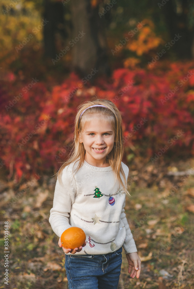 Wall mural Portrait of small happy girl holding an orange fruit and smiling in the park in warm autumn day