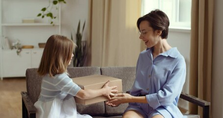 Young woman gives her daughter cardboard box with gift for birthday or holiday, congratulates child, little baby opens unpacks parcel, rejoices at new thing, measures clothes gratefully kisses mother - Powered by Adobe
