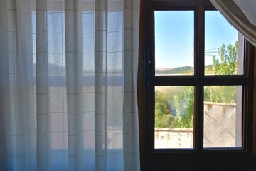 Wooden window with white curtain and views of houses, trees and sky.