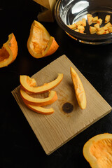 Slices of chopped pumpkin on wooden kitchen board on black table. Autumn seasonal vegetables cooking. Healthy eating habits, organic vegetarian food concept.