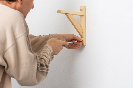 A man hangs a shelf in an apartment on a white wall, a worker fixes screws on a shelf corner with a screwdriver