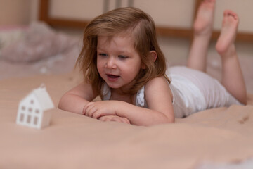 Fototapeta premium A little joyful girl with blond hair lies on the bed on a beige blanket. Baby on the bed. Happy childhood concept.