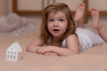 Fototapeta premium A little joyful girl with blond hair lies on the bed on a beige blanket. Baby on the bed. Happy childhood concept.