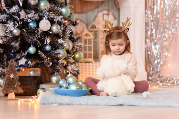 Merry Christmas and Happy Holidays! A cheerful girl with a rim of deer horns sits on a blanket next to a New Year tree on which blue balls hang.