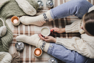 Young woman sits on plaid in cozy knitted woolen white sweater and socks holds cup of cocoa in her...