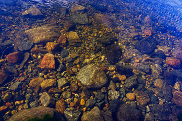 Stones at the bottom of a clear lake with clear water with a blue sky reflection.