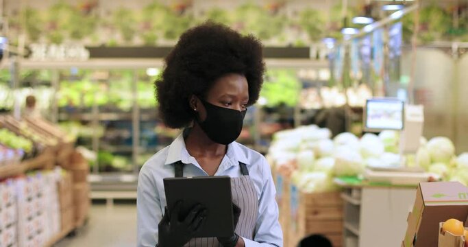 Close up portrait of pretty African Americn female manager in mask and gloves walking in supermarket and tapping on tablet. Beautiful woman employee typing on device in food store. Job concept