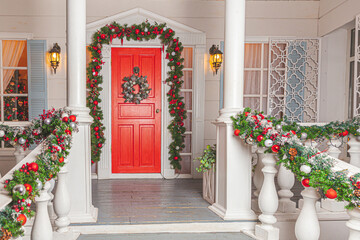 Fototapeta na wymiar Christmas porch decoration idea. House entrance with red door decorated for holidays. Red and green wreath garland of fir tree branches and lights on railing. Christmas eve at home