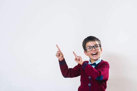 A portrait of a schoolboy with glasses and a red sweater is standing pointing fingers at a free space in the photo and smiling emotionally.