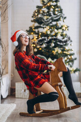 Pretty woman in shirt and socks have fun riding on the wooden swing horse toy, new year tree behind.