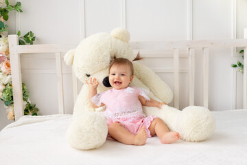 happy little baby girl six months old sitting on a white bed in pink clothes, with a big Teddy bear and smiling