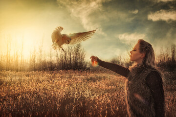 A wild owl preparing to land on the arm of a blonde woman, in front of a grass landscape