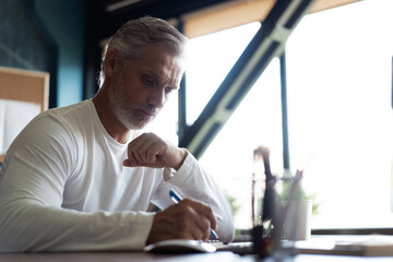 Casual Grey-haired businessman looking through paper documents. Male accounting manager reviewing financial report, sitting at table alone at office.
