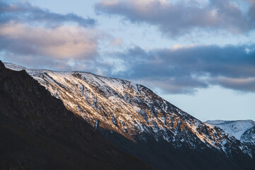 Atmospheric alpine landscape to snowy mountain ridge in sunset. Snow shines in golden light on mountain peak. Beautiful shiny snowy top. Evening cloudy sky. Wonderful scenery in gold sunset shades.