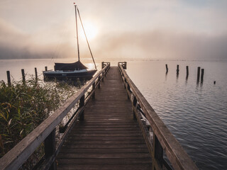 Fototapeta premium wooden jetty with a sailboat in the water of a lake in fog at sunrise