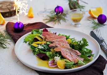 Warm salad with a mixture of salad leaves, baked duck and orange on a clay plate on a light concrete background in the New Year or Christmas style.